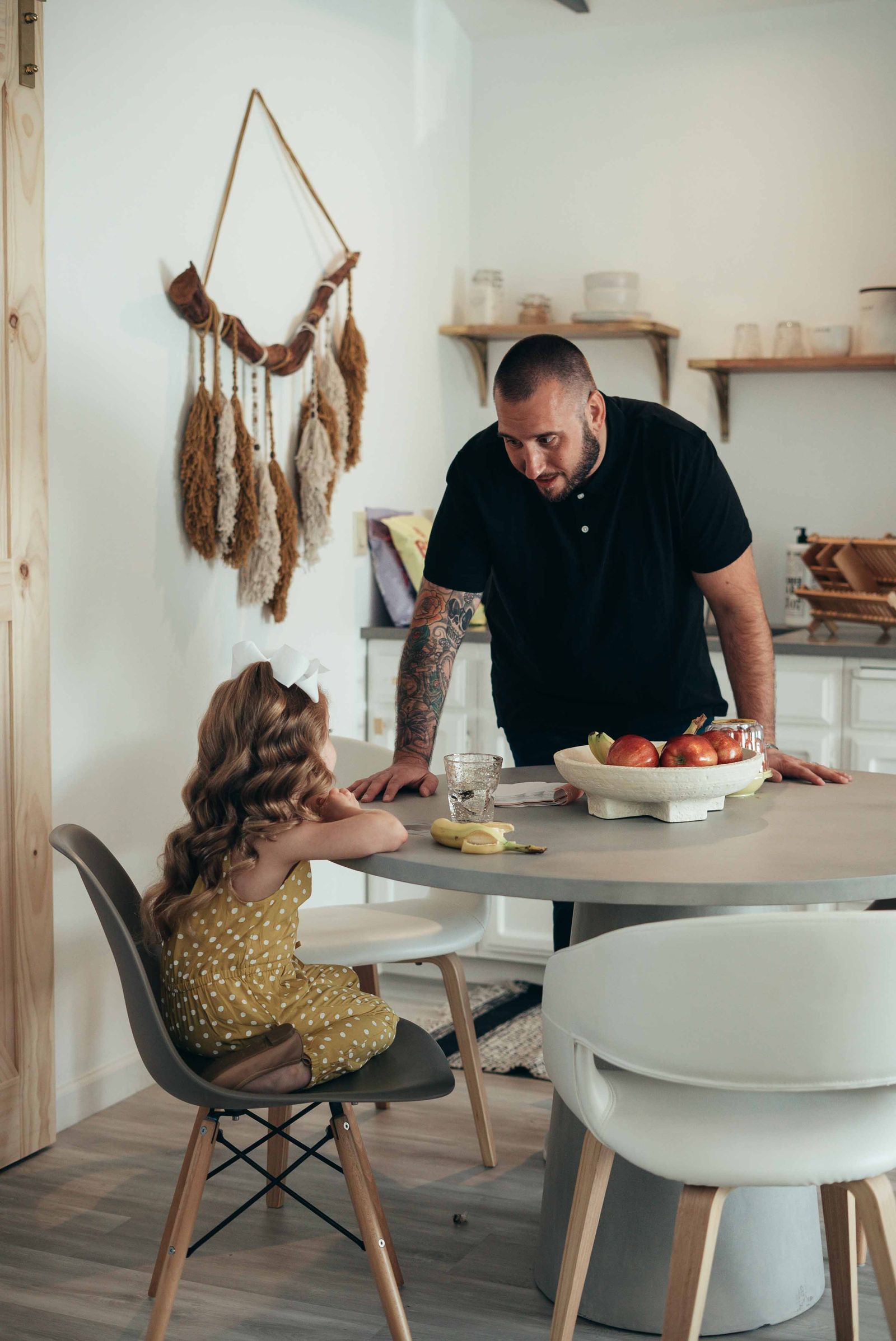 A father with tattooed arms leans on the kitchen table, chatting with his young daughter at breakfast