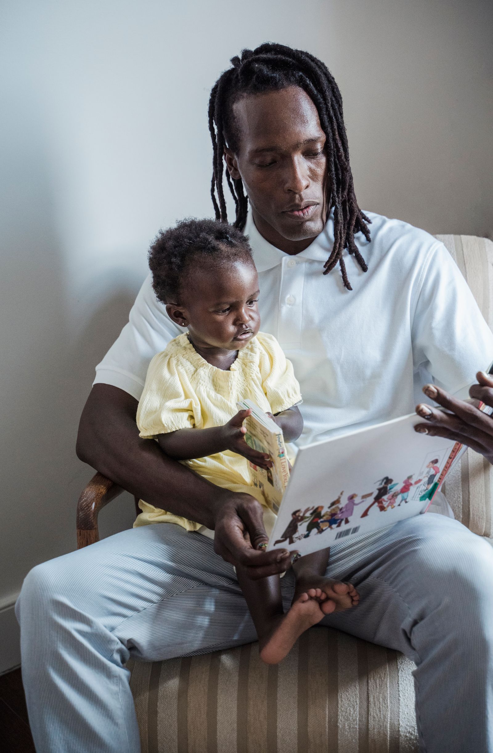 A father with long locs reads a picture book with his baby daughter on his lap
